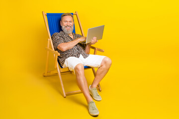 Full size photo of cheerful aged man sit lounger use wireless netbook isolated on yellow color background