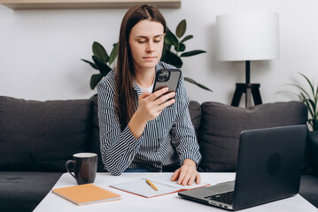 Selective focus of pensive young female using cellphone at workplace, sitting at laptop, chatting online, browsing Internet, shopping with ecommerce app. Communication gadgets and startup concept