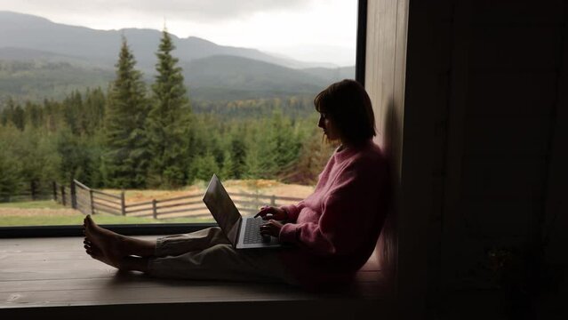 Woman works on laptop while sitting on a window with great view on mountains. Remote work and escape to nature concept