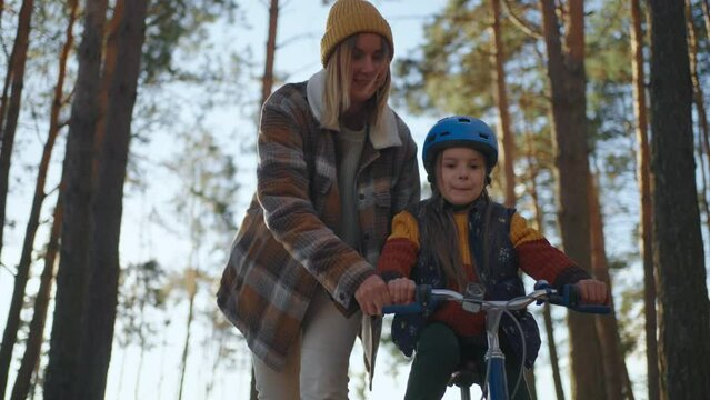 Happy mother having fun with her daughter who is learning yo ride bicycle in the park. Mother teaching her daughter to ride bicycle in park on sunny day