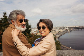 happy senior woman in stylish sunglasses hugging bearded husband in beige coat.