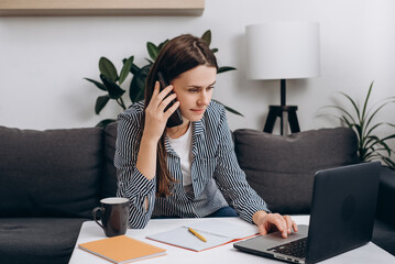 Attractive smiling young caucasian female sitting on cozy sofa talking on phone with client looking...