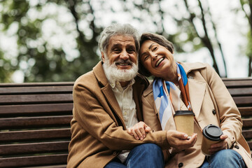 happy senior couple in coats holding paper cups with coffee to go and sitting on bench in park.