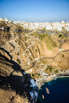 Elevated view of a steep cliff, hilltop town and a harbour on the water with a zigzag path up the cliffside on a Greek island.