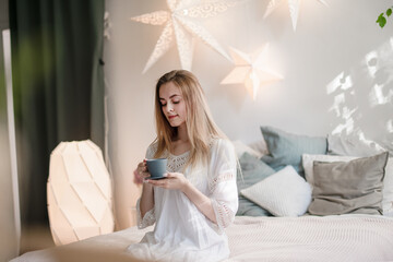 A happy young woman with a cup of coffee or tea in bed in a home bedroom is sitting on the bed. Slow life.
