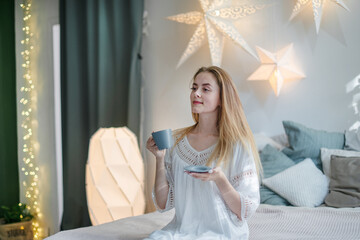 A happy young woman with a cup of coffee or tea in bed in a home bedroom is sitting on the bed. Slow life.