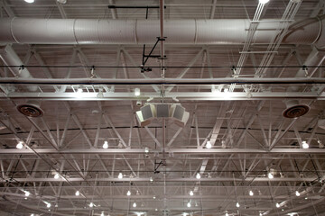 The ceiling of a large building, with air ducts and pipes, the rafters, gantry and struts, sound system speakers, lights and extractor units.