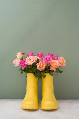 Bouquet of roses in yellow rubber boots on a gray table and a green background. Vertical frame.
