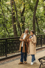 Fototapeta premium full length of cheerful senior couple in beige coats holding paper cups and walking in park.