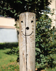Cute smiley face carved in wood pole outside in a playground. Happy and funny face carving as decoration on fence post. Lucerne Switzerland. Selective focus with defocused plants and building.