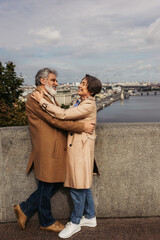 happy and senior couple in beige coats hugging on bridge near river.