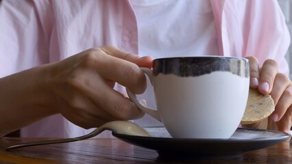 Close-up, unrecognizable woman drinking coffee with a cookie. Consumption of coffee and sweets during the coffee break. Coffee is addictive and affects the nervous system and circulation.