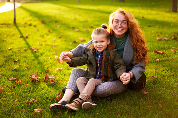 Naklejka na ściany i meble Smiling redhead woman and little boy sitting on grass lawn in city park on warm autumn day. Young mom hugs her son, they have fun and look at camera on sunny fall day. Foliage on green lawn.