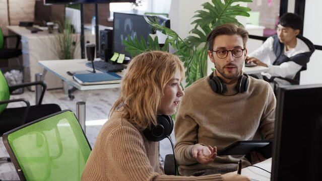 European woman and Caucasian male artist discuss the 3D model in a computer game and application development studio among other team members.
