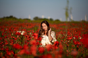 Fototapeta na wymiar artistic photo of a beautiful long haired woman in a classic white dress enjoying the sunset over a vast field of red flowers; a magical rural landscape during sunset