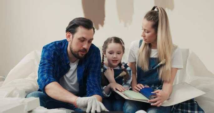 Happy family sitting on sofa in room where renovation work is taking place. Charming blonde woman helps husband daughter choose best shade of paint color to paint walls in living room.