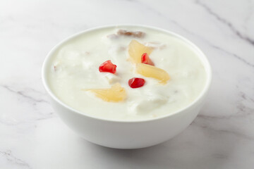 Close-up of Fruits raita or Yogurt & Pineapple or Ananas Raita garnished with pineapple piece and pomegranate, served in a white ceramic bowl over whiet granite background