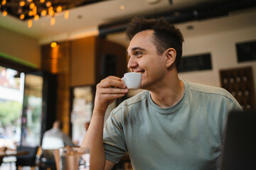 A young guy using his computer in cafe and talking on the phone while drinking water and coffee 