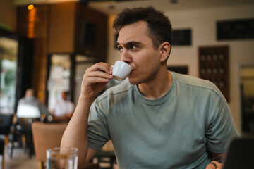 A young guy using his computer in cafe and talking on the phone while drinking water and coffee 