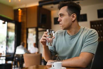 A young guy using his computer in cafe and talking on the phone while drinking water and coffee 