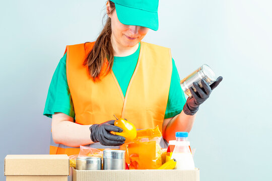 Free Food Distribution. Volunteer sorting food donation box. Young smiling woman wearing uniform cap and t-shirt, orange vest. Girl collects grocery sets, helping in-need people