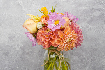 Close up of a flower arrangement placed in a glass jar. Peach and pink flowers. Dahlias, lisianthus, gomphrena, and celosia flowers. Gray background. Space for text.