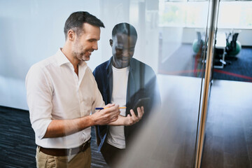 Two coworkers planning business ideas at office standing at glass wall looking at phone