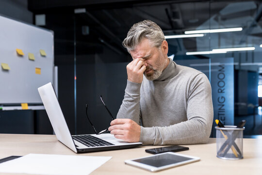 Stressful, tense, tired handsome male businessman sitting in office at desk with laptop and phone. Takes a break, took off his glasses, covered his eyes with his hand.