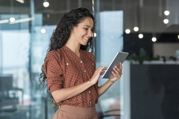 Young beautiful hispanic businesswoman standing in the office in the corridor. He holds a tablet in his hands, uses it, types, smiles.