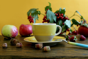  Autumn tea party: a yellow cup with hot tea, a spoon nearby, nuts, apples, a bouquet of viburnum on a wooden table, a yellow background