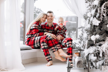 Fototapeta na wymiar Young happy family in the same pyjamas: smiling mom, dad, baby boy are sitting on big window by the festive decoration Christmas tree. The theme of the family holiday is New year and Christmas.