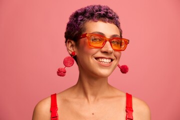 Fashion portrait of a woman with a short haircut in colored sunglasses with unusual accessories with earrings smiling on a pink bright background