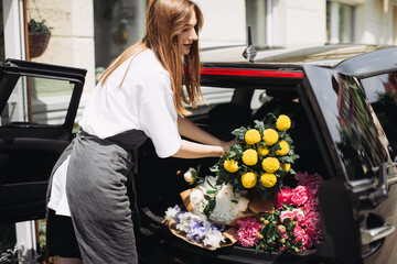 A woman florist puts flowers in the trunk of a car. Flower delivery concept. - Powered by Adobe