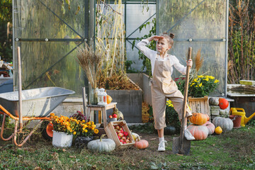 The girl harvests and stands near the greenhouse with a pitchfork.