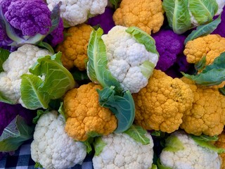 Whole heads of white, orange, and purple raw cauliflower with green leaves piled on tabletop at farmers market 