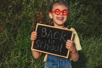 Portrait of happy little girl in red fake glasses keeps black chalk board with handwritten text...