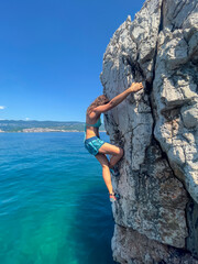 CLOSE UP: Female climber deep water solo climbing above clear blue seawater