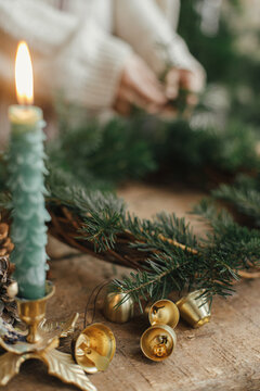 Golden bells on background of stylish christmas candle as fir tree and woman making christmas wreath on rustic wooden table. Holiday advent. Making Christmas rustic wreath, moody image