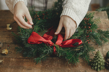 Woman hands decorating christmas wreath with red ribbon on rustic wooden table with pine cones and candles. Making Christmas wreath, moody holiday image. Holiday workshop, winter decor