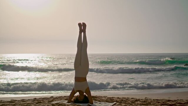 Woman doing headstand yoga pose on seashore at sunrise. Girl training balance.