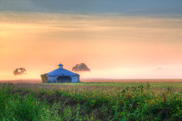 Fototapeta premium Barn between the trees Highway 94 in Warren County at dawn, the colorful sunlight dances on the low lying fog. 