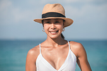 Portrait of young woman with straw hat standing at beach. Young smiling woman on vacation enjoy sea breeze and looking at camera.