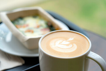 Latte art coffee in white mug with soft focus breakfast in background