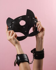 Women's hands in leather handcuffs holding black cat mask on pink background. BDSM accessories, domination, sex games © splitov27
