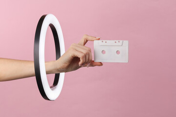 Woman's hand holds audio cassette through led ring lamp on pink background. Creative idea