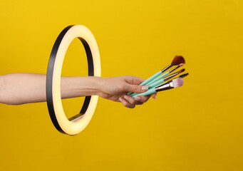 Woman's hand holds makeup brushes through led ring lamp on yellow background. Creative idea. Beauty...