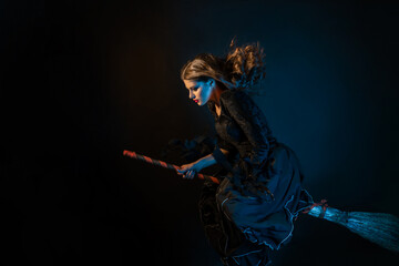 A young witch is rushing to the sabbath. A young beautiful brunette in a witch dress is flying on a broom, black background, photo in motion, grease
