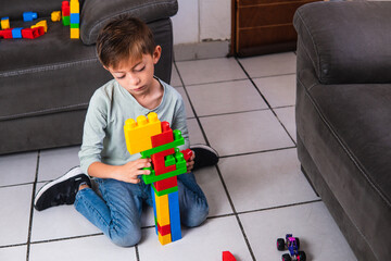 High quality photography. Boy playing with colored bricks. Boy playing with didactic toy on a messy room. Caucasian and blond boy playing in his living room next to the couch. - obrazy, fototapety, plakaty