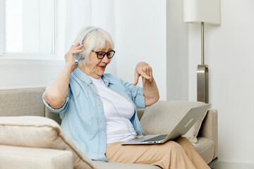 a sweet, happy elderly woman is sitting at home on a cozy sofa with headphones on her head and looking at a laptop monitor learning new things