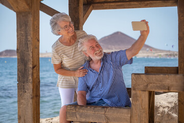 Happy caucasian senior couple sitting in the shade of the gazebo in front of the sea looking at mobile phone for a selfie - elderly pensioners enjoying sea vacation in a windy sunny day